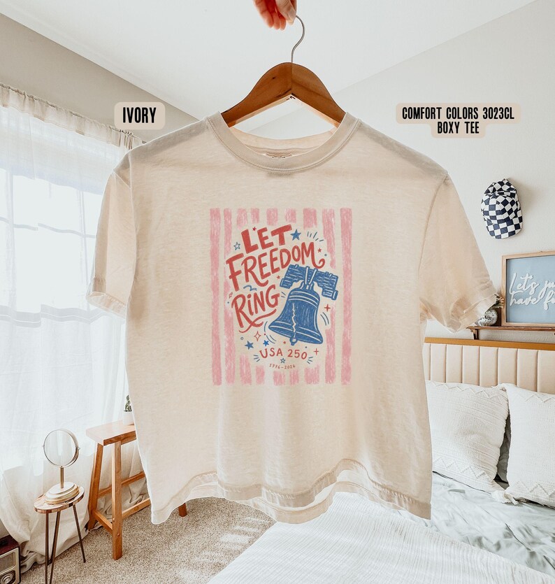 K&ouml;nnte beinhalten: Elfenbeinfarbenes Comfort Colors Boxy-T-Shirt mit der Aufschrift "Let Freedom Ring" und einer blauen Liberty Bell-Illustration. Die Grafik enth&auml;lt auch den Text "USA 250" und ist von rot-wei&szlig;en Streifen umrahmt.