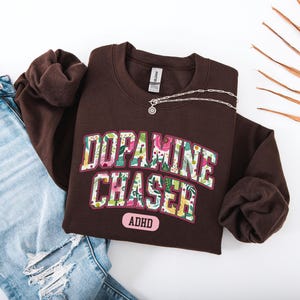 Pode incluir: Um moletom marrom escuro com o texto "Dopamine Chaser ADHD" em um design floral. Um colar de prata com um pingente est&aacute; no moletom. Jeans azuis claros s&atilde;o vis&iacute;veis no lado esquerdo da imagem.