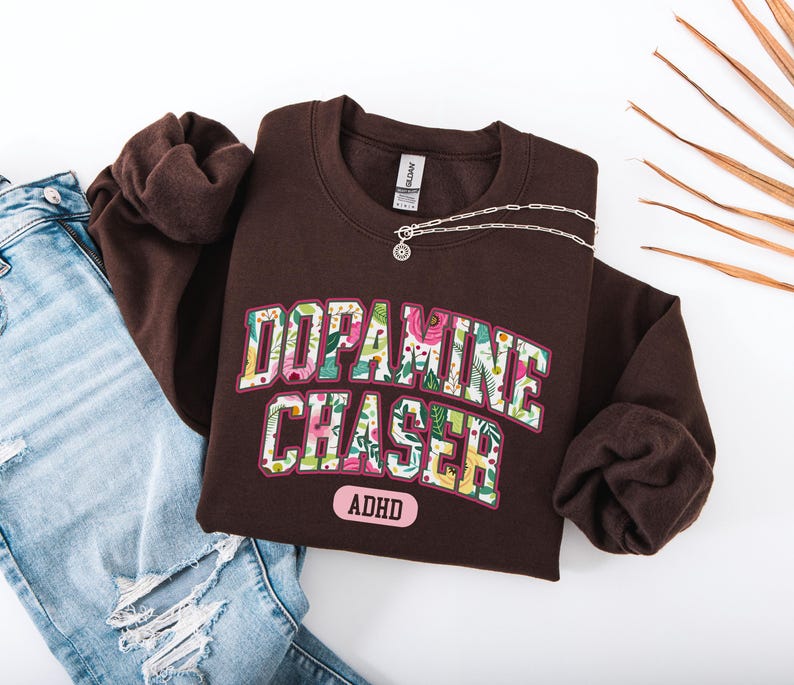 Pode incluir: Um moletom marrom escuro com o texto "Dopamine Chaser ADHD" em um design floral. Um colar de prata com um pingente est&aacute; no moletom. Jeans azuis claros s&atilde;o vis&iacute;veis no lado esquerdo da imagem.