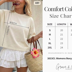 May include: A woman wearing an ivory-colored boxy tee shirt and white skirt, holding a straw tote bag with a pink flower. The image includes a size chart with measurements in inches for the tee shirt, ranging from S to 2XL.