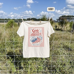 May include: Ivory-colored t-shirt with a patriotic design. The graphic features an eagle holding a waving American flag, with the words "GOD BLESS AMERICA" and "EST 1776" in a red and blue color scheme. The shirt is hanging on a clothesline.