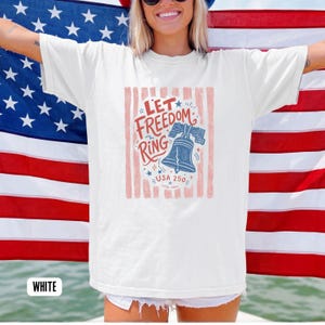K&ouml;nnte beinhalten: Wei&szlig;es T-Shirt mit einem patriotischen Design mit dem Text "Let Freedom Ring" und einer blauen Glockenillustration. Das Design enth&auml;lt rote und blaue Akzente, Sterne und den Text "USA 250". Das T-Shirt wird vor einer amerikanischen Flagge getragen.