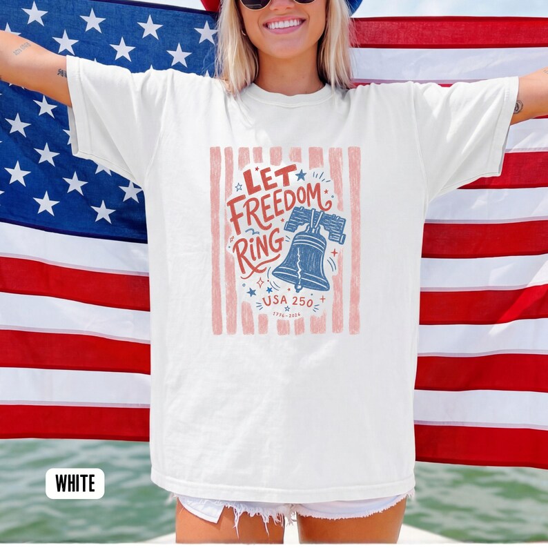 K&ouml;nnte beinhalten: Wei&szlig;es T-Shirt mit einem patriotischen Design mit dem Text "Let Freedom Ring" und einer blauen Glockenillustration. Das Design enth&auml;lt rote und blaue Akzente, Sterne und den Text "USA 250". Das T-Shirt wird vor einer amerikanischen Flagge getragen.