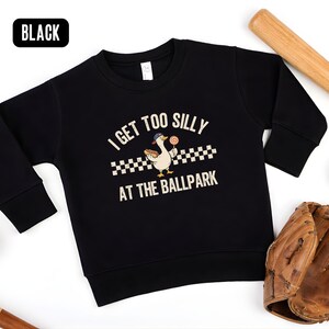 May include: Black sweatshirt with the text "I GET TOO SILLY AT THE BALLPARK" and a cartoon duck wearing a baseball cap and holding a lollipop. Baseball bats, baseballs, and a baseball glove are arranged around the sweatshirt.