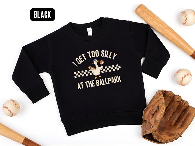 May include: Black sweatshirt with the text "I GET TOO SILLY AT THE BALLPARK" and a cartoon duck wearing a baseball cap and holding a lollipop. Baseball bats, baseballs, and a baseball glove are arranged around the sweatshirt.