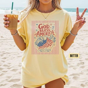 May include: Butter-colored t-shirt with a patriotic graphic. The design features an eagle holding a waving American flag, with the words "God Bless America" and "Est 1776". The shirt is a relaxed fit.