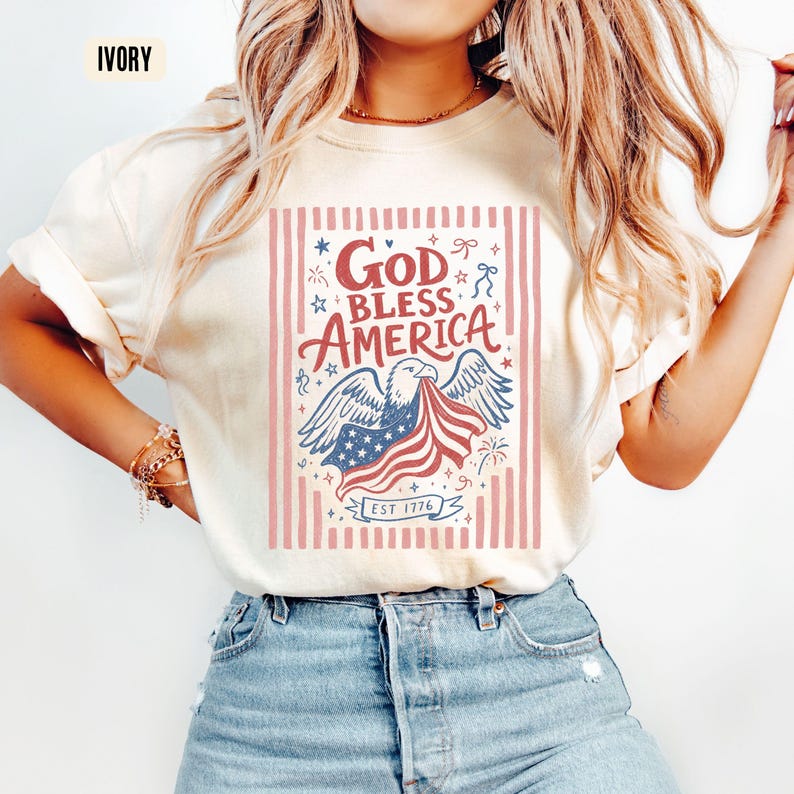 May include: Ivory-colored t-shirt featuring a patriotic design. The graphic includes an eagle holding a waving American flag, with the words "God Bless America" and "Est. 1776". The design is set against a backdrop of red and white stripes.