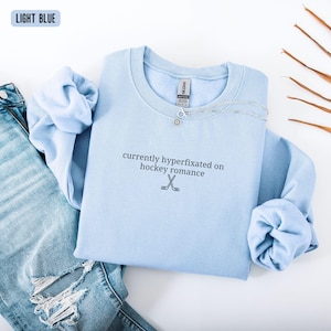 May include: A light blue crewneck sweatshirt featuring the black embroidered text currently hyperfixated on hockey romance and a small graphic of two crossed hockey sticks. The garment includes a Gildan brand label at the inner neckline.