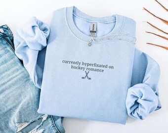 May include: A light blue crewneck sweatshirt featuring the black embroidered text currently hyperfixated on hockey romance and a small graphic of two crossed hockey sticks. The garment includes a Gildan brand label at the inner neckline.