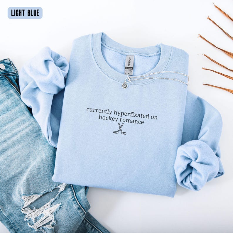 May include: A light blue crewneck sweatshirt featuring the black embroidered text currently hyperfixated on hockey romance and a small graphic of two crossed hockey sticks. The garment includes a Gildan brand label at the inner neckline.
