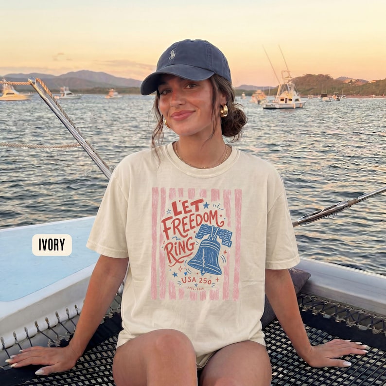 K&ouml;nnte beinhalten: Elfenbeinfarbenes T-Shirt mit dem Text "Let Freedom Ring" und einer blauen Glockengrafik. Das Design umfasst rote und rosa Streifen, Sterne und den Text "USA 250". Das Shirt wird auf einem Boot mit einem malerischen Hintergrund getragen.