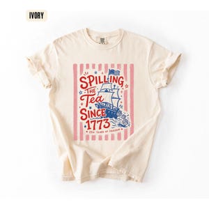 May include: Ivory-colored t-shirt with a graphic design. The design features a ship, stars, and the text "Spilling the Tea Since 1773" in red and blue, set against a pink striped background. The shirt also includes the text "250 Years of Freedom".
