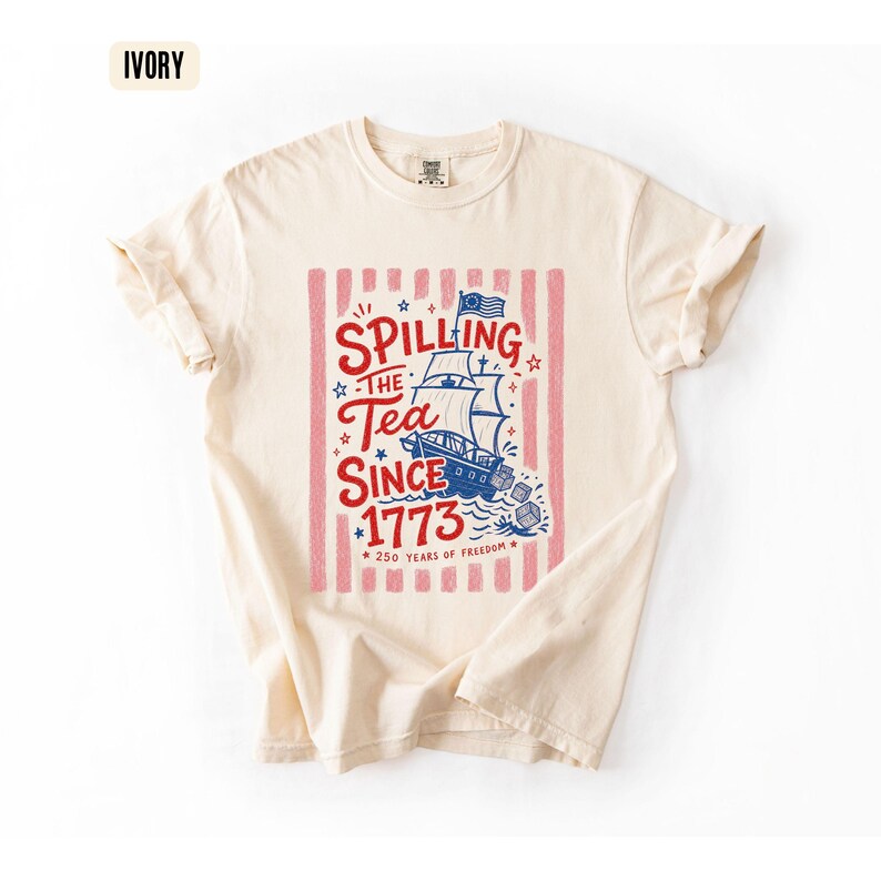 May include: Ivory-colored t-shirt with a graphic design. The design features a ship, stars, and the text "Spilling the Tea Since 1773" in red and blue, set against a pink striped background. The shirt also includes the text "250 Years of Freedom".