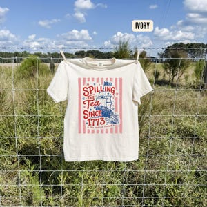 May include: Ivory-colored t-shirt with a patriotic design. The graphic features a ship, stars, and the text "Spilling the Tea Since 1773" in red and blue, with "250 Years of Freedom" below. The shirt is hanging on a clothesline.