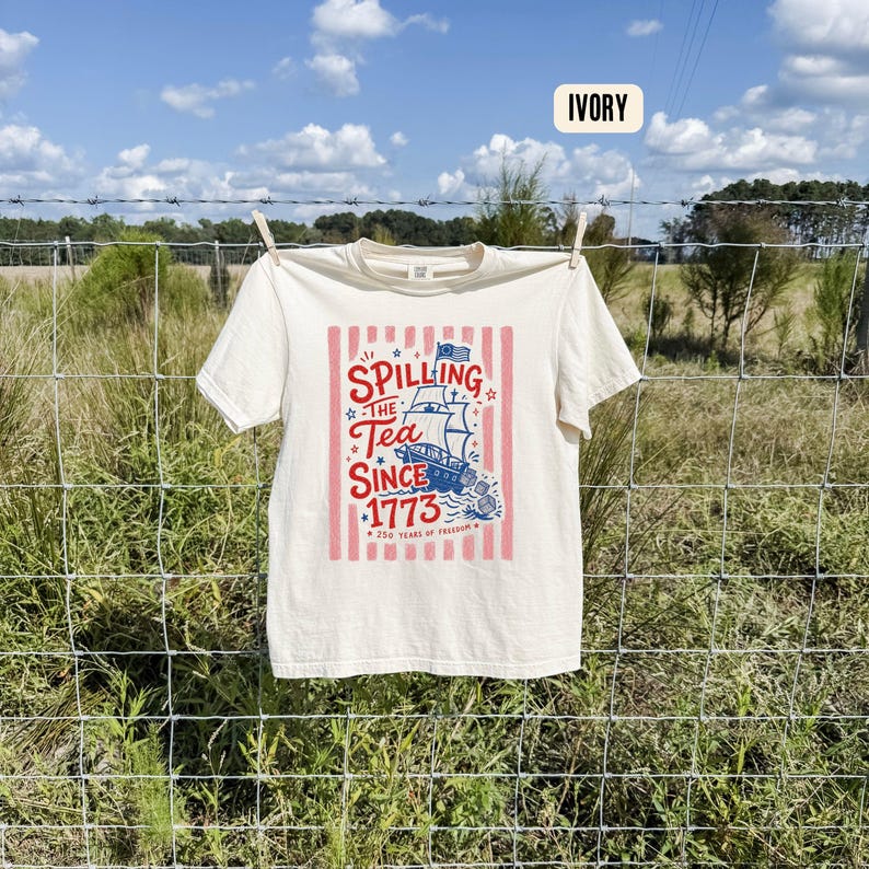 May include: Ivory-colored t-shirt with a patriotic design. The graphic features a ship, stars, and the text "Spilling the Tea Since 1773" in red and blue, with "250 Years of Freedom" below. The shirt is hanging on a clothesline.
