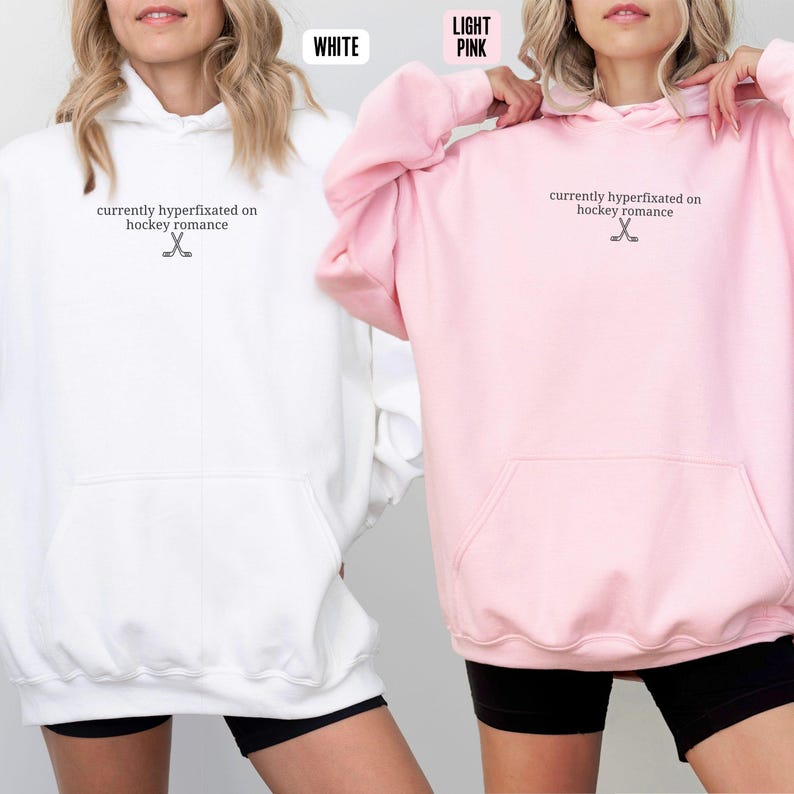 May include: A white hoodie and a light pink hoodie, both featuring a front kangaroo pocket and the black text "currently hyperfixated on hockey romance" accompanied by a small graphic of two crossed hockey sticks centered on the chest.