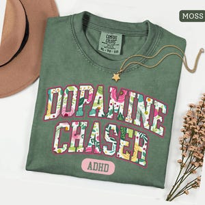 Pode incluir: Uma camiseta verde musgo com as palavras "Dopamine Chaser" em um design floral e a palavra "ADHD" abaixo. Um colar com pingente de estrela dourada e um chap&eacute;u marrom est&atilde;o ao lado da camisa.