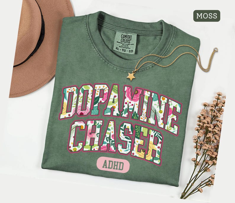 Pode incluir: Uma camiseta verde musgo com as palavras "Dopamine Chaser" em um design floral e a palavra "ADHD" abaixo. Um colar com pingente de estrela dourada e um chap&eacute;u marrom est&atilde;o ao lado da camisa.