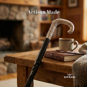 May include: A black walking cane with a silver handle featuring an ornate leaf design. The cane is resting on a wooden table, with the text "Artisan Made" visible in the background. A leather-bound journal and a mug are also on the table.