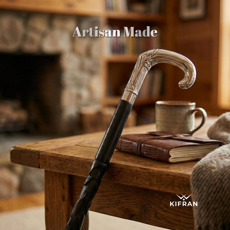 May include: A black walking cane with a silver handle featuring an ornate leaf design. The cane is resting on a wooden table, with the text "Artisan Made" visible in the background. A leather-bound journal and a mug are also on the table.