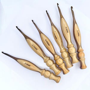 May include: Six wooden crochet hooks with varying sizes, from 2.5MM to 5.0MM, are arranged diagonally. Each hook features a light-colored wood handle with a darker wood accent and the size printed on the handle.