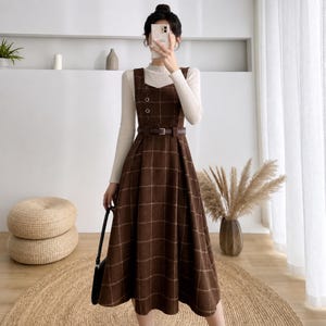 May include: A brown plaid pinafore dress with a sweetheart neckline, two decorative buttons on the bodice, and a matching brown belt at the waist, worn over a long-sleeved white mock neck top. The dress features a full skirt and a classic grid pattern.