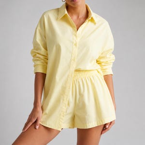 May include: A light yellow button-down shirt with a classic collar and long sleeves paired with matching light yellow shorts featuring an elasticated waistband. The set is made of a solid-coloured fabric with a smooth texture and a relaxed fit.