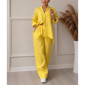 Op de afbeelding: Een felgele tweedelige set bestaande uit een overhemd met lange mouwen, knoopsluiting, kraag en een borstzak, gecombineerd met een bijpassende broek met hoge taille en wijde pijpen. De stof heeft een zichtbaar geweven textuur over het kledingstuk.