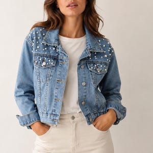 May include: A medium-wash blue denim jacket featuring a classic collar, long sleeves, and button-front closure. The upper front panels and collar are embellished with scattered white faux pearls of varying sizes. It is worn over a plain white shirt.