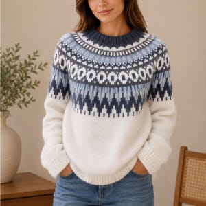 May include: A long-sleeve knit sweater featuring a white base and a yoke decorated with a blue and dark gray geometric fair isle pattern. The garment has a ribbed crew neckline, ribbed cuffs, and a ribbed hem, providing a structured fit.