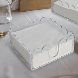 May include: Clear acrylic napkin holder with a scalloped edge design. The holder contains a stack of white napkins. The holder has a curved handle on the side for easy access. The design is modern and minimalist.