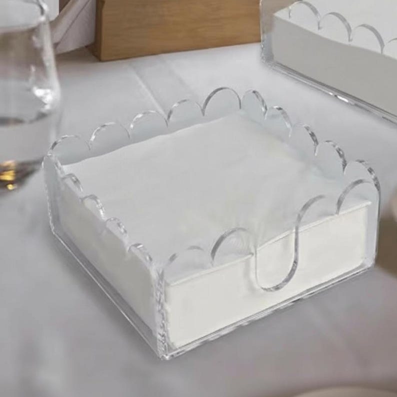 May include: Clear acrylic napkin holder with a scalloped edge design. The holder contains a stack of white napkins. The holder has a curved handle on the side for easy access. The design is modern and minimalist.