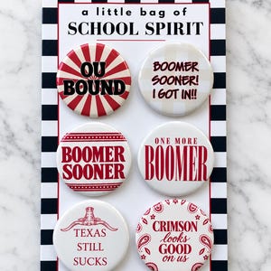 May include: A set of six circular pin-back buttons on a rectangular card with a black and white checkered border. The card reads a little bag of SCHOOL SPIRIT. The buttons feature red and white designs with text including OU BOUND, BOOMER SOONER! I GOT IN!!, BOOMER SOONER, ONE MORE BOOMER, TEXAS STILL SUCKS, and CRIMSON looks GOOD on us.