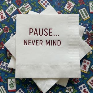 May include: A stack of square white paper napkins with a textured border. The top napkin features the dark red printed text "PAUSE... NEVER MIND" centered in a clean, sans-serif font. The napkins are layered to show their square shape.