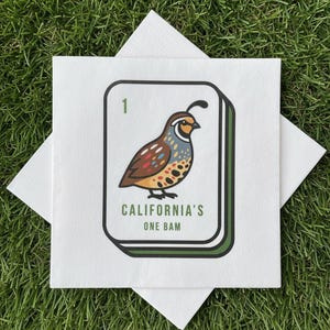 May include: White square napkin featuring a quail bird illustration, with text reading "CALIFORNIA'S ONE BAM". The bird is depicted in brown, tan, and blue. The napkin rests on green grass.