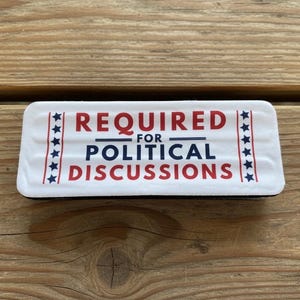 May include: A rectangular white magnet with rounded corners featuring the red text REQUIRED and POLITICAL DISCUSSIONS, with the blue text FOR in between. Vertical borders on each side display a column of blue stars on a white background.