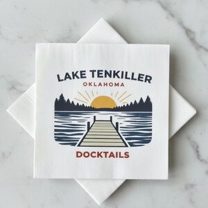 May include: White square cocktail napkins with a graphic design. The design features a lake scene with a dock, trees, and a rising sun. The text reads "Lake Tenkiller Oklahoma" and "Docktails" in a dark blue and red font.