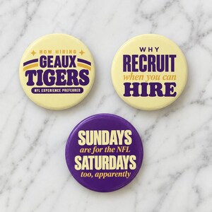 May include: Three circular pinback buttons. The top left is pale yellow with purple text reading NOW HIRING GEAUX TIGERS NFL EXPERIENCE PREFERRED. The top right is pale yellow with purple text reading WHY RECRUIT when you can HIRE. The bottom is purple with yellow text reading SUNDAYS are for the NFL SATURDAYS too, apparently.