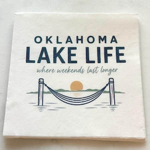 May include: A square white paper napkin featuring the dark blue text OKLAHOMA LAKE LIFE and the script text where weekends last longer. Below the text is a graphic of a hammock between two posts with a yellow sun over green hills.