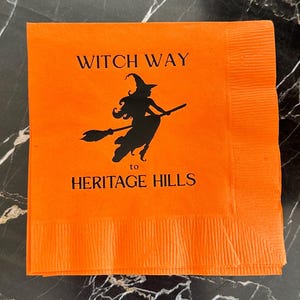 May include: Orange Halloween-themed paper napkin with the text "WITCH WAY to HERITAGE HILLS" and a black silhouette of a witch flying on a broom. The napkin is folded and has a textured edge.