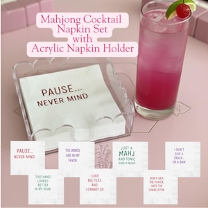 May include: A Mahjong Cocktail Napkin Set with an acrylic napkin holder. The set includes white napkins with phrases like "PAUSE... NEVER MIND" and "JUST A MAHJ AND TONIC KIND OF NIGHT". A pink cocktail with a cherry garnish is also in the image.