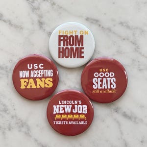 May include: Four circular pinback buttons with text. One white button reads FIGHT ON FROM HOME. Three maroon buttons read USC NOW ACCEPTING FANS, USC GOOD SEATS still available, and LINCOLN'S NEW JOB TICKETS AVAILABLE with a graphic of five stadium seats.