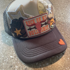 May include: A brown and beige trucker cap with a mesh back. The hat has patches including a rose, stars, and a patch that reads "BACK ON MY BULL SHIT" with a bull skull. A small orange heart is on the brim.