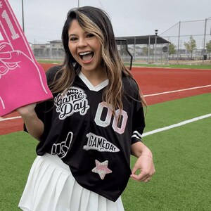 May include: A black jersey with white accents and the words "Game Day" and "00" in white and pink. The jersey is paired with a white pleated skirt. A pink foam hand is held up in the air.