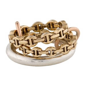 May include: A collection of mixed metal rings. The rings include a silver band, a gold chain-link ring, and two rose gold rings. The rings are intertwined, creating a layered effect. The rings are likely made of precious metals.