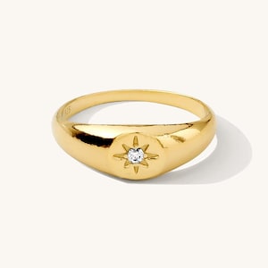 Peut inclure: Une bague dorée avec un motif d'éclat d'étoile et une petite pierre précieuse transparente au centre. La bague a une surface lisse et polie et un anneau arrondi. La bague est de taille 9.25.