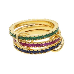 Op de afbeelding: Goudkleurige ringen met gekleurde edelstenen. De ringen zijn op elkaar gestapeld, met groene, magenta en blauwe stenen. De ringen zijn verbonden door gouden lussen.