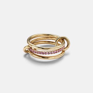 May include: A gold ring set with multiple bands, one of which is set with small pink gemstones. The ring has a unique design with interlocking circles and a connecting loop, creating a modern and elegant look.