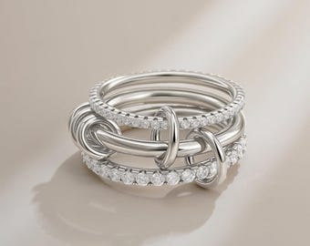 May include: A silver ring set featuring multiple bands intertwined with connecting loops. Some bands are encrusted with small, sparkling gemstones. The design is modern and elegant, with a polished finish.