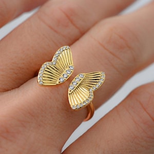 Puede incluir: Un anillo dorado con diseño de alas de mariposa. Cada ala está adornada con pequeñas gemas transparentes en los bordes y tiene un patrón detallado en forma de abanico. El anillo se muestra en un dedo.